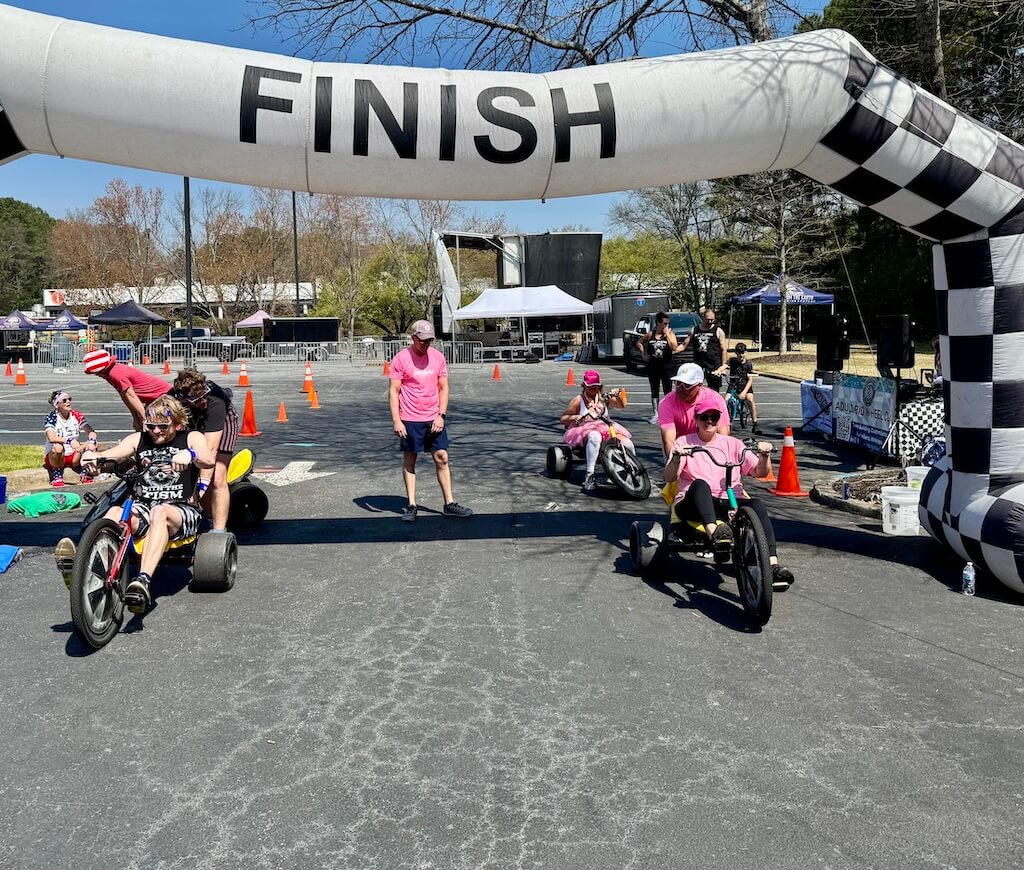 People on tricycles crossing a finish line in an outdoor race, with an inflatable arch and traffic cones marking the path.