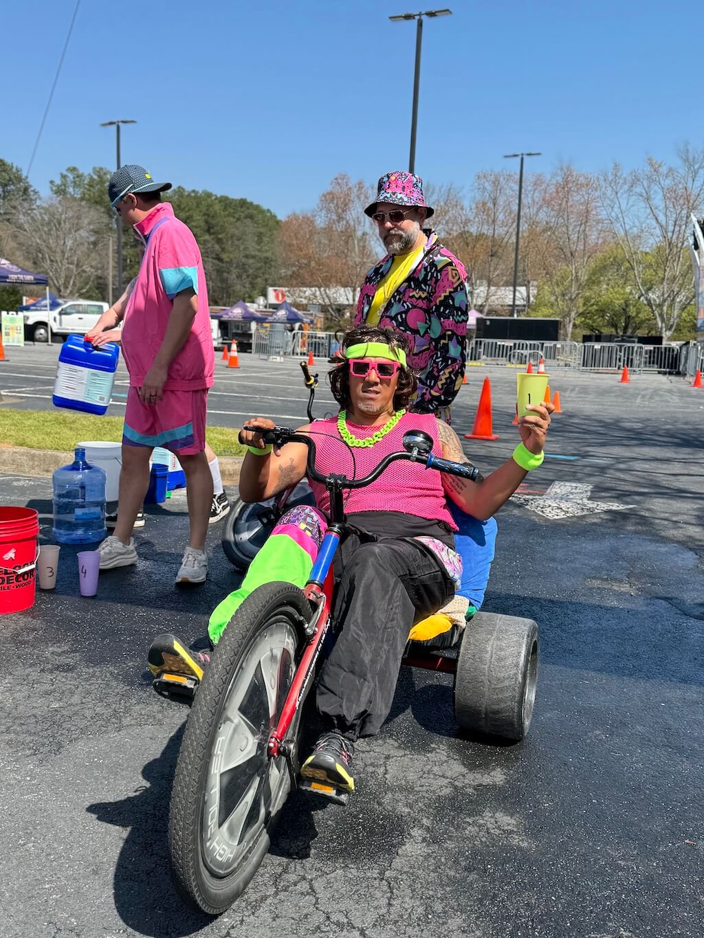 Person in colorful attire rides a tricycle, holding a cup. Two others in colorful outfits stand nearby with water jugs, outdoors on a sunny day.